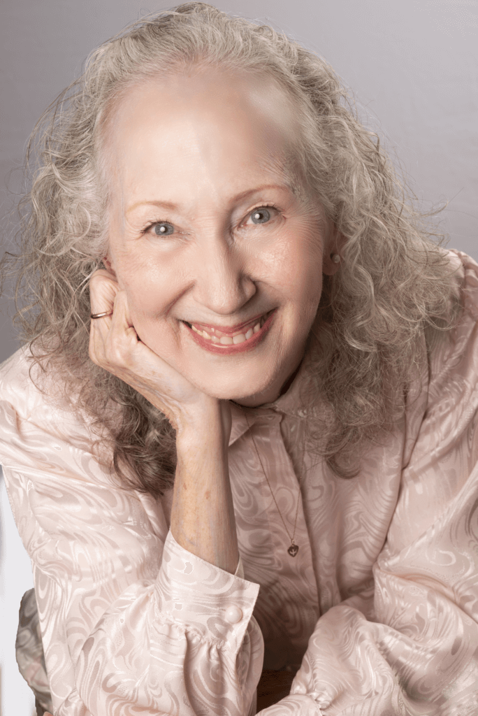 Elderly woman with gray curly hair smiling, wearing a light patterned blouse, and resting her chin on her hand.