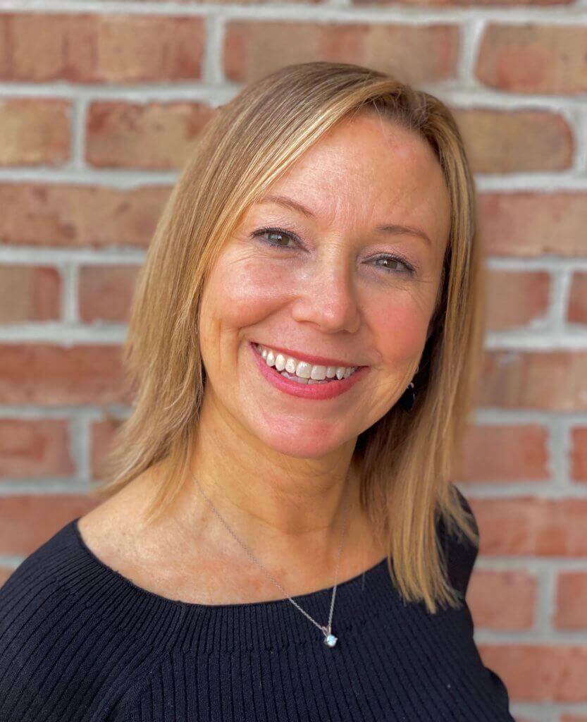 A smiling woman against a brick wall, possibly discussing visual supports for sensory diets and self-regulation in kids.